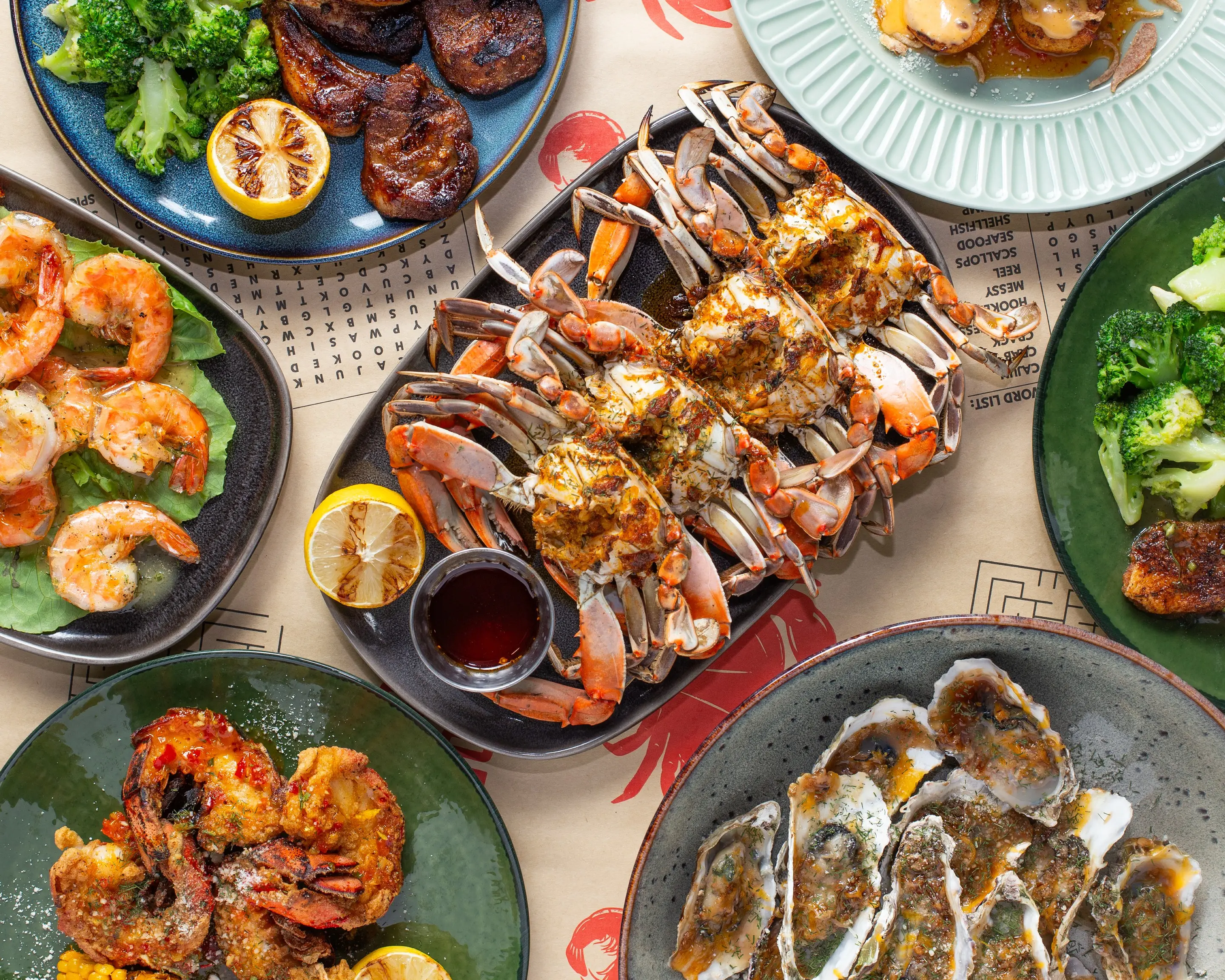 A full table photographed from above on the restaurant’s printed kraft paper: a black platter of seasoned blue crab at the centre with a cup of dipping butter, ringed by grilled lamb chops with broccoli, poached shrimp on lettuce, chargrilled oysters, scallops and charred lemon halves.